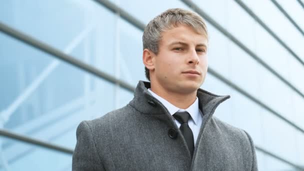 Hombre guapo, retrato. Primer plano de joven profesional en traje y abrigo mirando a la cámara, de pie en un edificio de vidrio de oficina ligero y moderno — Vídeos de Stock