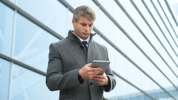 Retrato de hombre de negocios en traje y abrigo sosteniendo la tableta. Hombre usando tableta pantalla táctil al aire libre, sobre fondo moderno edificio de vidrio de oficina — Vídeos de Stock