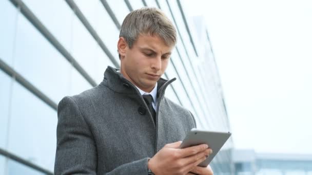 Primer plano retrato de hombre de negocios en traje y abrigo que sostiene la tableta. Hombre usando tableta pantalla táctil al aire libre, sobre fondo moderno edificio de vidrio de oficina — Vídeos de Stock