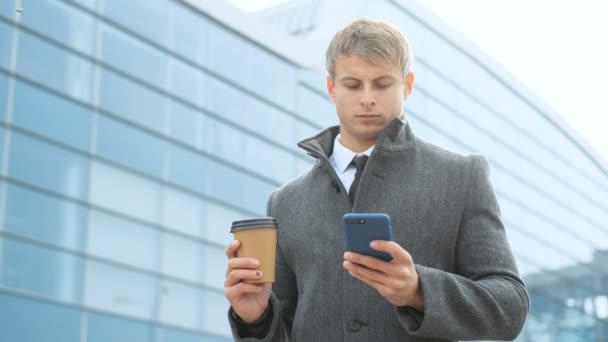 Ernstige zakenman tekstberichten op een mobiele telefoon buiten. Man met pak en de jas, staande in de buurt van moderne glas kantoor gebouw en holding een kop koffie. Bedrijfsconcept. — Stockvideo