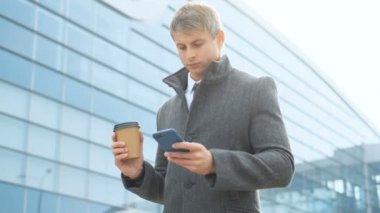 Ciddi iş adamı manifatura açık bir mobil cep telefonuyla portresi. Takım elbise ve ceket giyen adam modern cam ofis kahve holding Binası yürüyor. Kentsel profesyonel