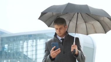Altında siyah şemsiye tutarak akıllı telefon, manifatura meslektaşım ve modern cam Ofis Binası yakınındaki yürürken bakarak adam portresi