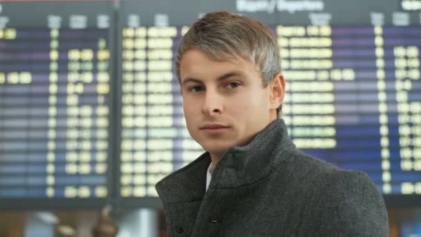 Close up portrait of businessman wearing suit and coat, looking at camera. Man checking his flight on the arrivals electronic table background. — Stock Video