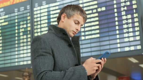 Portrait d'un homme d'affaires sérieux en costume et manteau utilisant un téléphone intelligent pour vérifier son vol sur le fond de la table électronique des arrivées . — Video