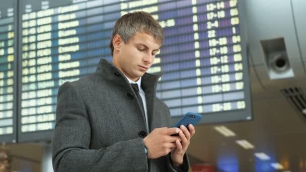 Nära upp skott av allvarlig man i moderna flygplatsterminalen och textning på smart telefon med vänner. Affärsman med smart telefon för att kontrollera sin flykt på ankomster elektroniska bordsbakgrund. — Stockvideo