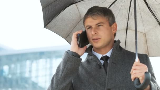 Man met paraplu en slimme telefoon lopen op straat. Portret van de knappe jongeman praten op smartphone, onder paraplu genieten van regenachtig weer — Stockvideo