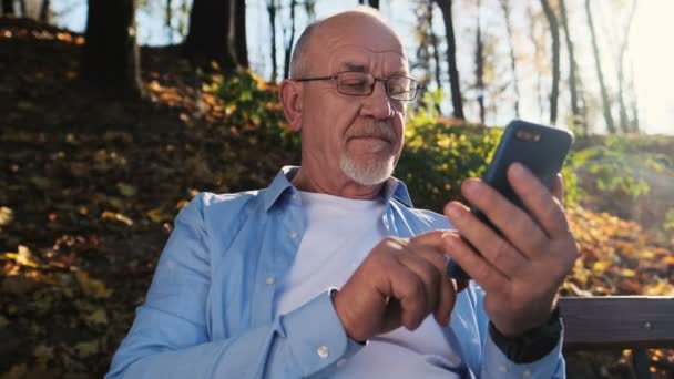 Hombre mayor usando el teléfono móvil, sosteniendo el teléfono inteligente, mensajes de texto, navegar por Internet, y el uso de la aplicación. Retrato de hombre con barba y gafas, se sienta en un banco del parque y usa un dispositivo — Vídeo de stock