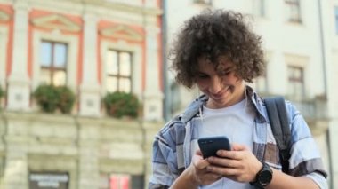 Portre çekici kıvırcık genç adam telefon kullanarak bir kadeh. Gülümseyen ve onun smartphone yakışıklı hipster dinleme müzik kulaklık, Avrupa'nın eski şehir merkezinin sokakta gülüyor