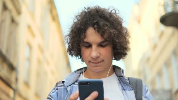 Ritratto ravvicinato di un bel ragazzo sorridente e ridente che ascolta musica dallo smartphone in cuffia, sulla strada del centro storico in Europa. Studente, concetto urbano . — Video Stock