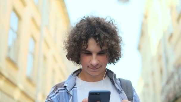 Крупный план портрета улыбающегося и смеющегося симпатичного кудрявого молодого человека, слушающего музыку со своего смартфона в наушниках, подростка, гуляющего по улице старого центра города в Европе — стоковое видео