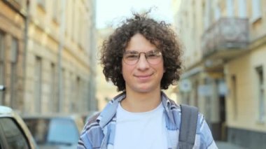 Rüzgar ve gözlük, kameraya bakarak üfleme kıvırcık saçlı genç gülümseyen adam portresi kapatın. Avrupa. Ağır çekim. Öğrenci, kentsel kavramı.