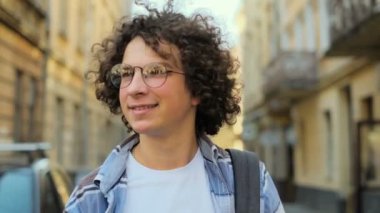 Esen rüzgar ve gözlük, kameraya bakarak, Avrupa'nın eski şehir merkezinin sokakta yürürken kıvırcık saçlı adam gülümseyen genç portresi kapatın. Ağır çekim. Öğrenci, kentsel kavramı.