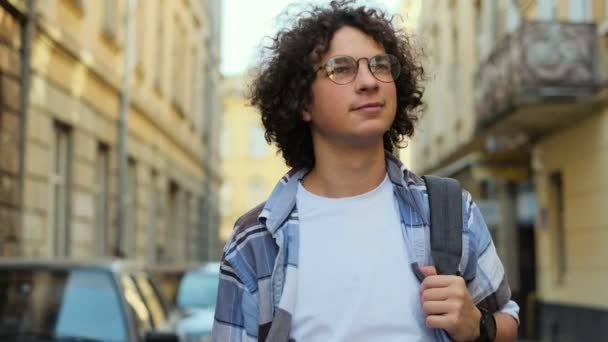 Primer plano retrato de un joven hipster sonriente con el pelo rizado soplando en el viento y las gafas, el hombre mirando a la cámara, caminando por la calle del centro de la ciudad vieja en Europa. A cámara lenta. Estudiante, concepto urbano . — Vídeos de Stock