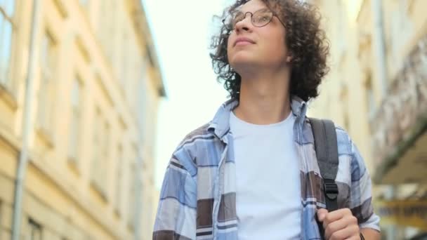 Portrett av smilende og leende, kjekk, krøllete ung mann med briller som går på gata i Europe ved sollys, mens han ser på kamera. Student, bykonsept . – stockvideo