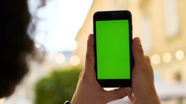 クロマ キー、新しい技術コンセプトの通り背景に緑色の画面で携帯電話のスマート フォンを保持している男。クロマ キー。屋外。男は路上に立っている間携帯電話を使用して。ショット バック ビュー. — ストック動画