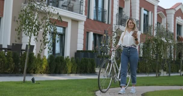 Élégante fille hipster avec vélo parlant sur le smartphone dans la rue de la vieille ville européenne. Heureuse belle fille utilisant le téléphone intelligent extérieur, portrait de fille d'été — Video