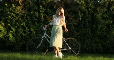 Bisikleti ile gülümseyen güzel genç kadın portre. Drees yeşil doğal arka plan üzerinde duran ve kameraya bakarak retro vintage Bisiklet ile güzel kız moda