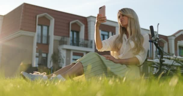 Hipster 女孩与自行车使用手机在智能手机应用程序上发短信在街上, 并采取自拍。女孩躺在绿草上, 在城市公园休息, 骑自行车躺在草坪上. — 图库视频影像