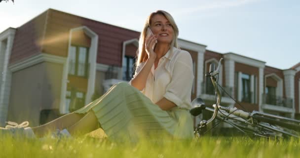 Charmante jonge meisje, praten over de telefoon, vrouw liggend op groen gras, rusten in stadspark, fiets liggend op gazon. — Stockvideo