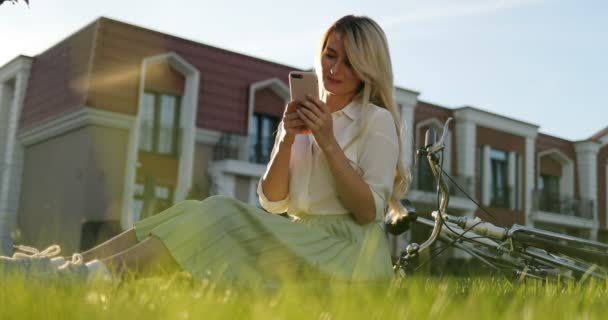 Giovane ciclista sorridente donna sdraiata su erba verde nel parco, utilizzando il telefono cellulare. Ragazza hipster con la bicicletta utilizzando un telefono sms su smartphone app in una strada — Video Stock