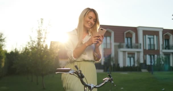 Smarphone を使用してレトロな自転車に乗って緑のドレスで幸せな少女の笑みを浮かべてください。アプリを使用して、電話番号、テキスト メッセージを屋外自転車と幸せの美しい女性の肖像画 — ストック動画