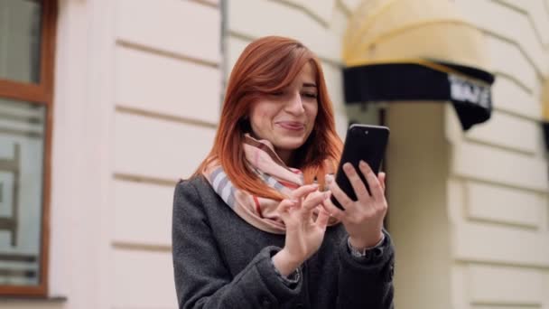 Close-up van aantrekkelijke glimlachend gember jonge vrouw op stedelijke street, met behulp van moderne smartphone buiten. Dame ontvangt korting voor winkelen, controle e-mail vak op smart phone — Stockvideo