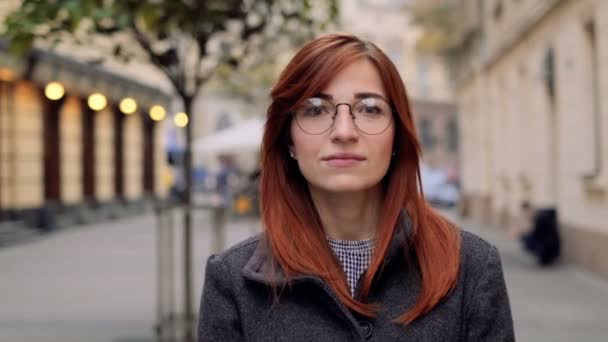 Primer plano retrato de mujer joven sonriendo con el pelo rojo en las gafas. Mujer atractiva sonriendo y mirando a la cámara en la ciudad serie de personas reales, confianza bastante . — Vídeo de stock