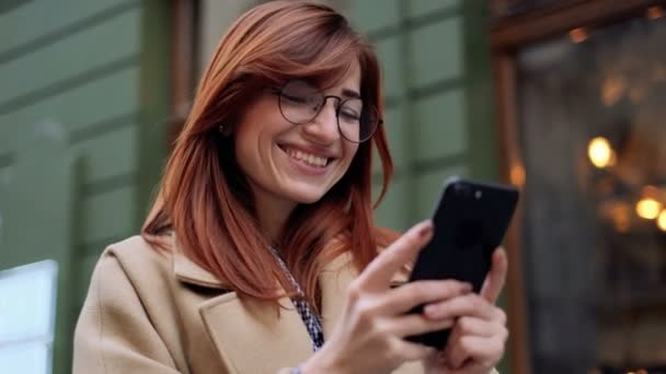 Mujer sonriente usando la aplicación en el teléfono móvil durante el paseo por las calles de la ciudad vieja. Mensajes de texto femeninos con teléfono móvil, escribiendo mensajes de texto en el teléfono celular. Primer plano retrato, estilo de vida, urbano, bokeh . — Vídeo de stock