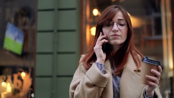 Close-up de mulher atraente em olhar elegante andando na rua urbana, usando telefone inteligente moderno e beber café para sair. Senhora de óculos falando no celular enquanto caminha na rua . — Vídeo de Stock