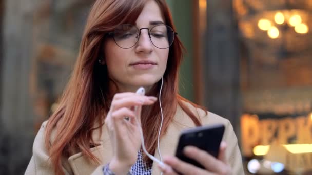 Porträtt av söt flicka i hörlurar och glasögon lyssnar på musik och surfa på mobiltelefon. City lights bakgrund. Kvinna som surfar på internet, rullar en sociala medier webbplats med smart telefon. — Stockvideo