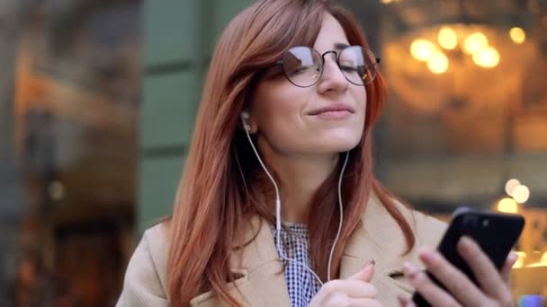 Retrato de menina bonito em fones de ouvido e óculos ouvindo música e navegação no telefone móvel ao ar livre. Luzes da cidade fundo. Mulher usando aplicativo, percorre um site de mídia social usando telefone inteligente . — Vídeo de Stock