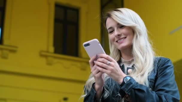 Mulher e usa smartphone ao ar livre. Muito hipster, estudante mensagens de texto menina e usando o aplicativo no telefone inteligente no fundo amarelo, olhando para a câmera e sorrir. Estilo de vida, urbano — Vídeo de Stock