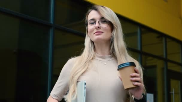 Primer plano de mujer atractiva en aspecto elegante caminando por la calle urbana, bebiendo café para salir al exterior. Señora en gafas que sostiene el ordenador portátil mientras camina por la calle . — Vídeo de stock