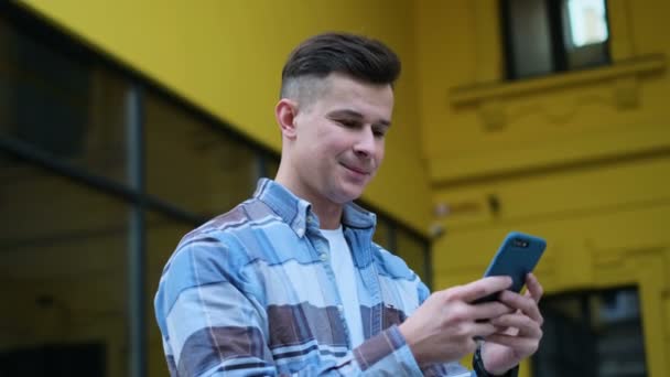 Sonriendo joven guapo usando sus rollos de teléfonos inteligentes a través de las redes sociales se alimentan en el dispositivo, risitas y sonrisas de noticias sobre aplicaciones en la calle del centro de la ciudad en Europa, fondo amarillo . — Vídeos de Stock