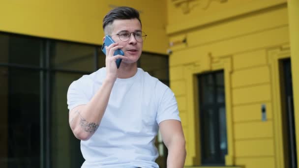 Обычный молодой человек, использующий смартфон. Портрет человека, говорящего на смартфоне, улыбающегося счастливо в белой футболке и очках на улице — стоковое видео