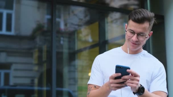 男子使用智能手机在社交媒体上滚动设备, 微笑着从街上的应用程序新闻。男人的肖像改变了手机上的歌曲和曲目, 在耳机里对音乐进行了磨写. — 图库视频影像
