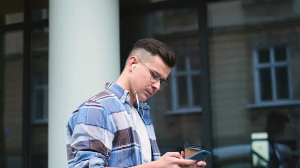 Portrét muže s smartphone pití kávy z jednorázových pohárek na městské ulici. Pohledný mladý muž komunikaci na smartphone s úsměvem jistý. — Stock video