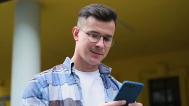 Yakın çekim portre yakışıklı genç adamın onun smartphone kullanarak aygıt, kıkırdıyor ve gülümsüyor uygulamaları Caddesi üzerinde şehir merkezi Avrupa'nın news feed sosyal medya üzerinden kaydırır,