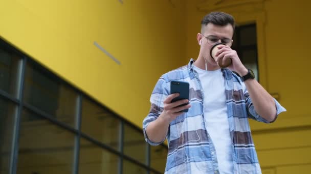 Ο άνθρωπος χρησιμοποιώντας το smartphone του κύλιση μέσω των social media που τρέφονται με τη συσκευή, γέλια και χαμόγελα από ειδήσεις σχετικά με τις αιτήσεις επί της οδού. Πορτρέτο του ο άνθρωπος που πίνει καφέ από διαθέσιμου χαρτί Κύπελλο — Αρχείο Βίντεο