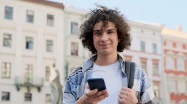 Yakışıklı genç adam onun smartphone kullanarak sosyal medya aygıtında besleme yoluyla verilirse, News uygulamalara ve kamera sırasında bakarak gülümser açık sokaklarda yürümek