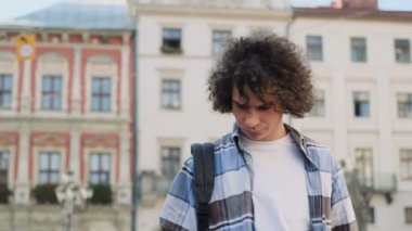 Sokakta yürüyen şık yakışıklı kıvırcık genç adam portresi. Hipster gülümseyen, blogger smarphone kullanarak, aygıtta yem ve kahve tutan sosyal medya aracılığıyla kayar
