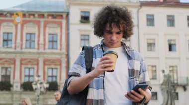 Şehir, gitmek için cep telefonunuzun mesaj yazarak ve kahve içme öğrenci, ağır çekim dışında yürürken modern akıllı telefon kullanarak şık beyaz kıvırcık yakışıklı genç portresi.