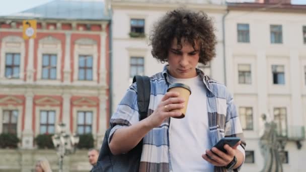 Portrait de élégant beau jeune homme bouclé caucasien utilisant un téléphone intelligent moderne tout en marchant dans la ville, étudiant tapant un message texte sur le téléphone portable et boire du café à emporter, ralenti à l'extérieur . — Video