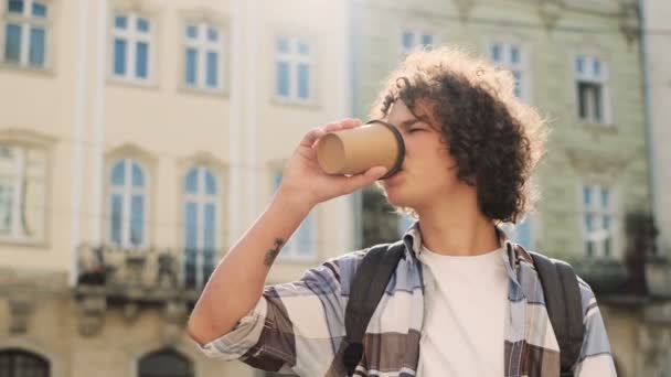 Portrét stylový pohledný kavkazské kudrnaté mladý muž chůzi ve městě, pít kávu s sebou, Zpomalený pohyb mimo. — Stock video