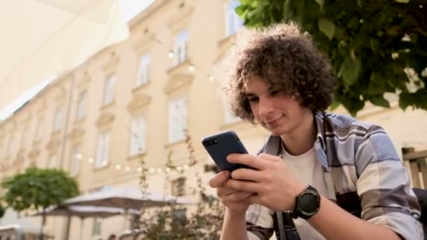 Pohledný mladý muž, turistické nebo student l v hipster oblečení, pomocí svého smartphonu svitky prostřednictvím sociálních médií se živí zařízení, kontrola mapy nebo čtení novinek na app, rozhlížel se kolem, venkovní — Stock video