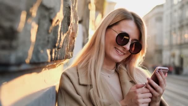 Retrato de mujer en ropa de otoño y gafas de sol escribiendo mensaje en su teléfono inteligente al aire libre. Chica usando gadget digital, se desplaza a través de las redes sociales en el dispositivo, leyendo noticias en la aplicación, al atardecer . — Vídeos de Stock