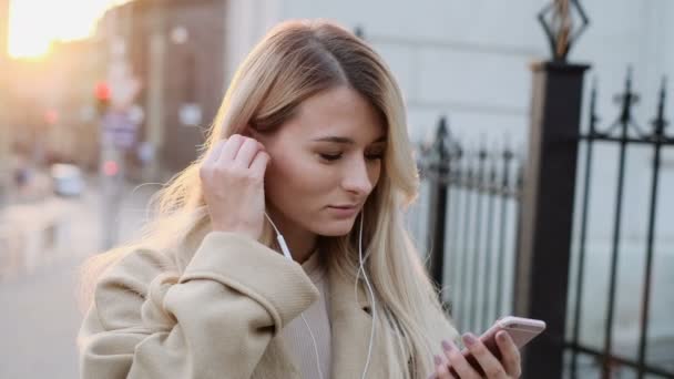 Menina sorridente em fones de ouvido usando smartphone, ouvir música e navegação no smartphone. Retrato ao ar livre. O blogueiro percorre as mídias sociais no dispositivo, alterando faixas e lendo notícias no aplicativo — Vídeo de Stock