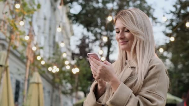 秋天衣服的女人的肖像写在她的智能手机户外的消息。女孩使用数字小玩意, 在设备上的社交媒体上滚动, 在应用上阅读新闻。. — 图库视频影像