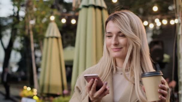 Retrato de elegante jovem bonito sorridente menina usando telefone inteligente moderno enquanto caminha na cidade. Lady digitar mensagem de texto no celular e beber café para ir, esperando amigo lá fora . — Vídeo de Stock