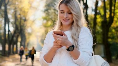 Kız beyaz kazaklı mesaj onun smartphone açık havada üzerinde yazma. Dijital gadget kullanarak kadın verilirse sosyal medya, günbatımında app, haber okuma, kameraya bakıyor.